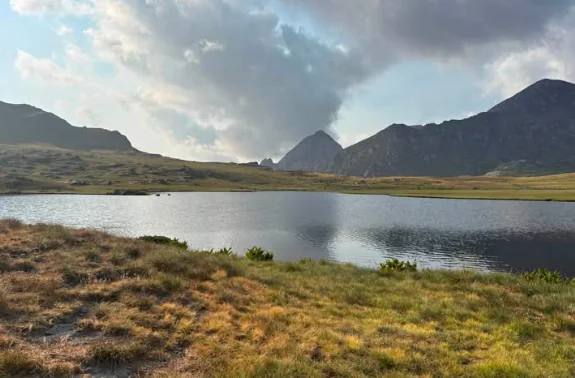 Klidné horské jezero mezi travnatými břehy a tmavými vrcholy pod mraky
